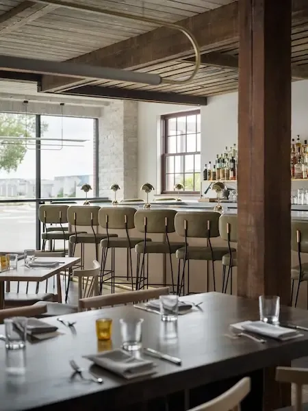 A modern, stylish restaurant interior designed by a Maine architect, featuring a wooden ceiling, green bar stools, neatly set tables, and large windows for natural light.