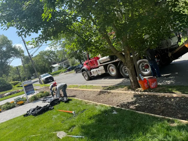 Workers install landscaping next to a residential street with a large truck. Green grass and trees frame the scene.