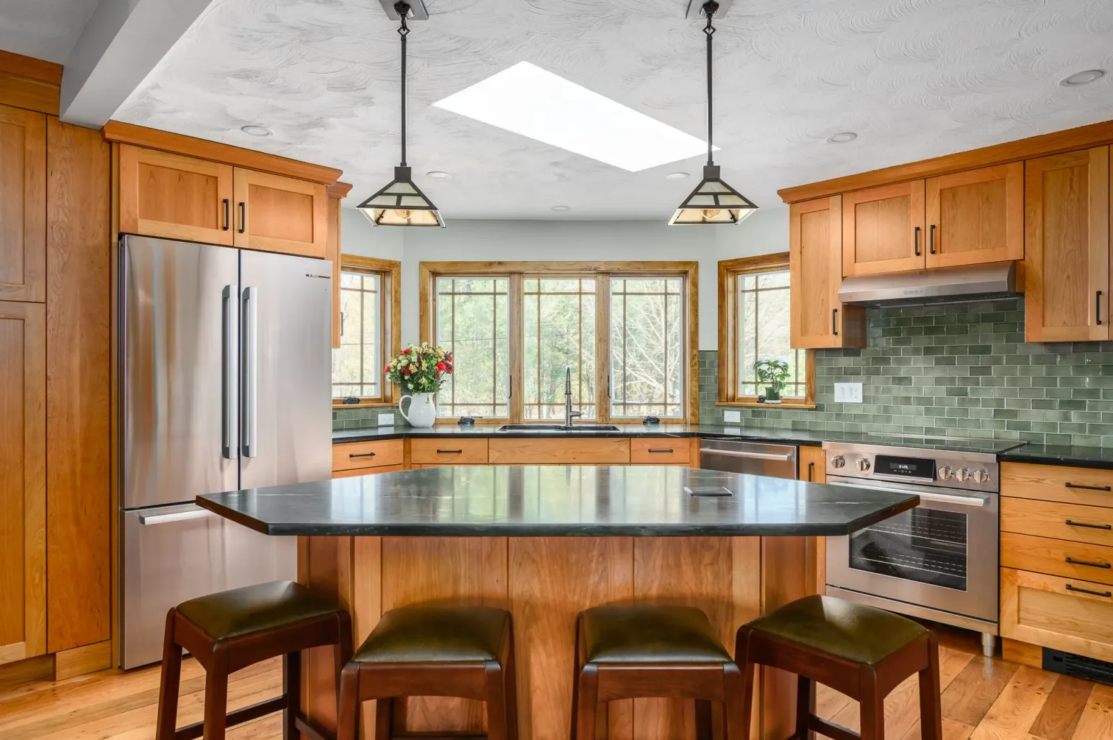 A kitchen with wood cabinets, green tile backsplash, stainless steel appliances, a black countertop island, and three chairs. There are windows with wood trim letting in natural light. Rooms With A View Tile