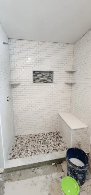 Bathroom shower with white subway tile walls, small built-in corner shelves, a small window with tile accents, a marble and pebble stone patterned shower floor, and construction supplies including a bucket of white tile grout and a green lid.