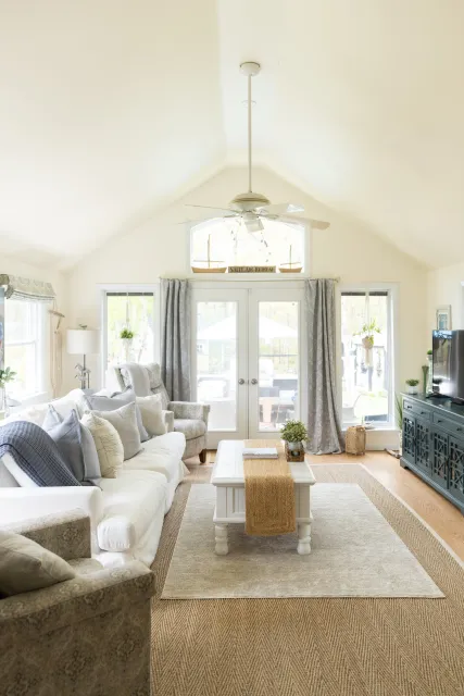 Living room with beige sofas, a white coffee table with a burlap runner, a gray sideboard, a ceiling fan with lights, large windows with curtains, and a carpeted floor.