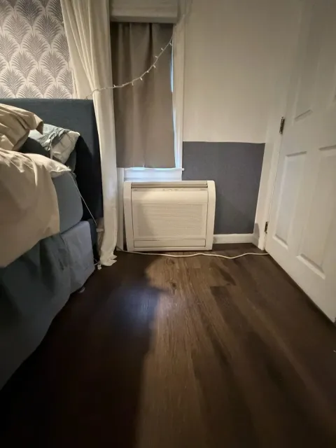 Small bedroom with bed, window and curtain, white door, and portable AC unit on the floor.
