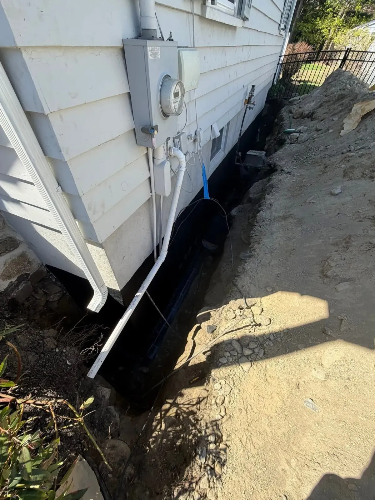 Damaged foundation along a house exterior, with black waterproofing and muddy ground beside it.