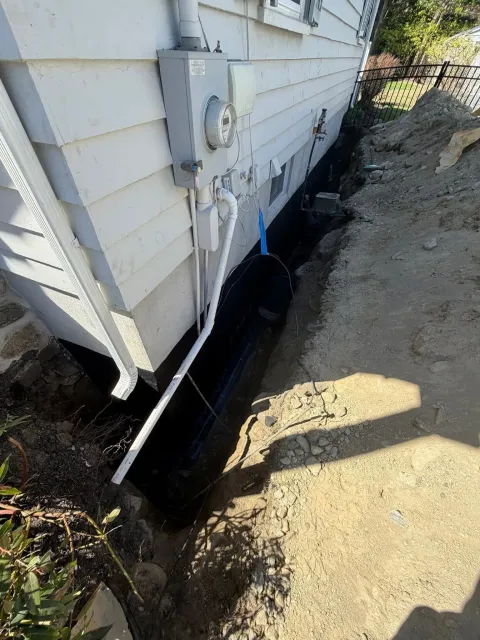 Damaged foundation along a house exterior, with black waterproofing and muddy ground beside it.