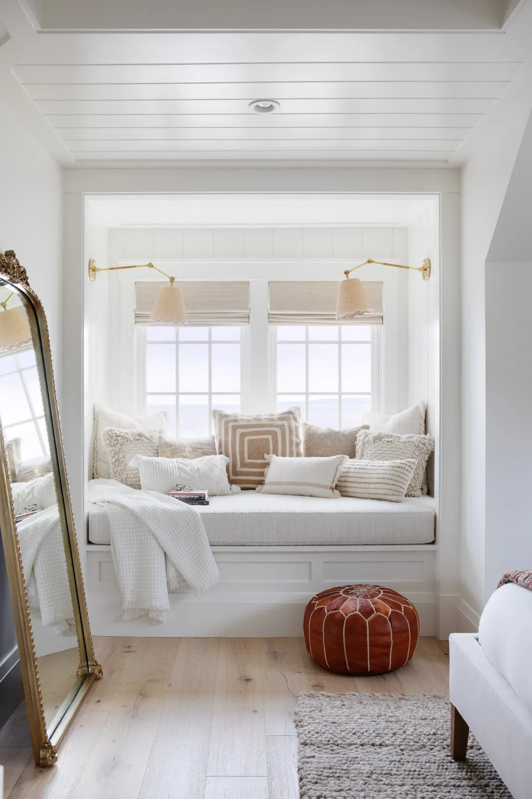 Hardwood floor refinishing in a Cohasset living space featuring a cozy window seat with beige and white pillows, two windows facing the ocean, a hanging lamp, a brown leather ottoman, a white armchair, a gold-framed mirror, and a rug on wood flooring