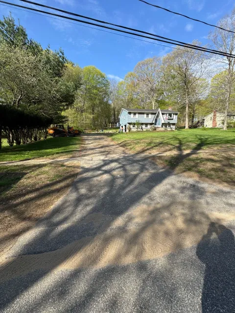 Gravel driveway leading to a white house under a blue sky, with trees and power lines overhead