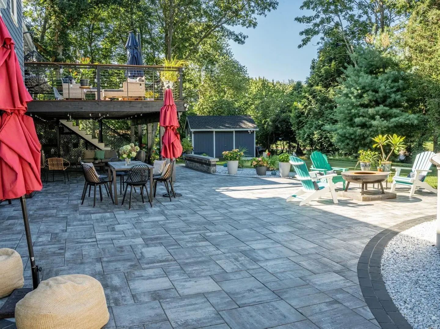 Patio with stone pavers, outdoor tables, red umbrellas, teal chairs, and lush trees in a sunny backyard