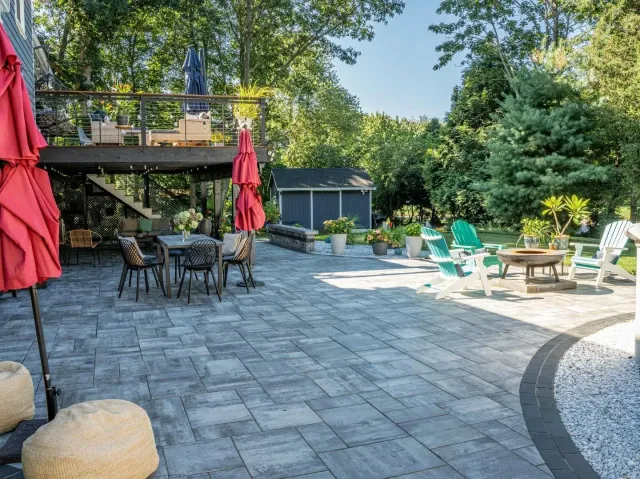 Patio with stone pavers, outdoor tables, red umbrellas, teal chairs, and lush trees in a sunny backyard
