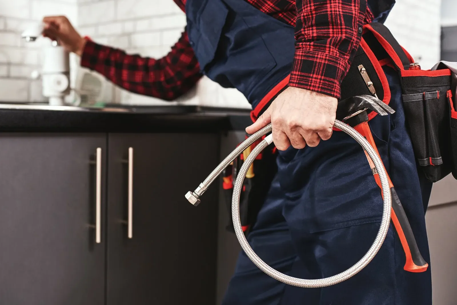 A plumber is holding a coiled metal hose and a wrench, working in a kitchen with black cabinets and a white brick wall background.