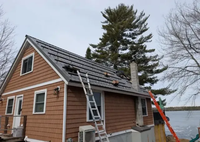 Workers installing solar panels on the roof of a lakeside wooden house with ladders leaning on it.