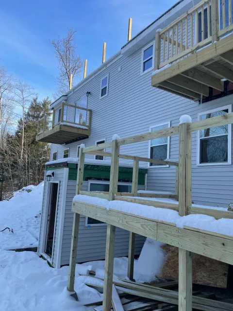 Exterior view of a building under construction with a wooden deck and snow around. The building has white vinyl siding, multiple windows, and small balconies, with photo taken during winter.