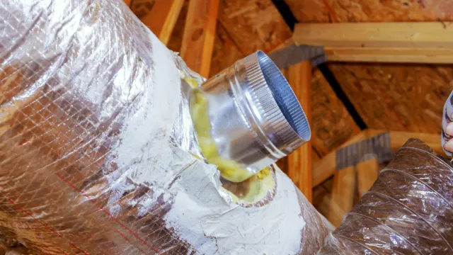 A hand holding a clear plastic cup over a cylindrical object wrapped in plastic, with a tube connecting the cup to the object in a wooden attic space.
