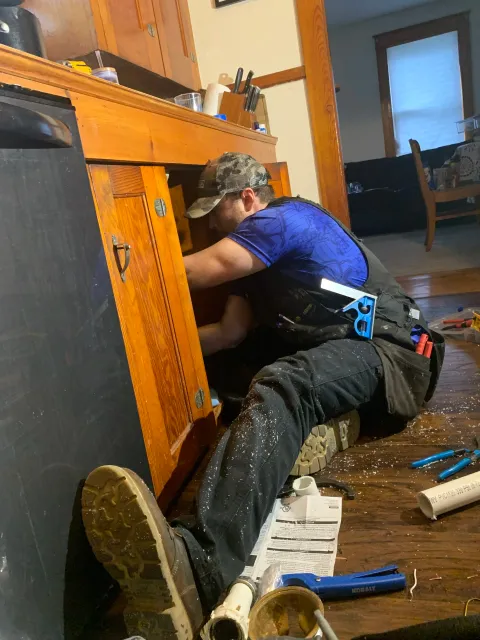 A man working on patching a wooden cabinet underneath a kitchen counter, surrounded by tools and pipes on a wooden floor.