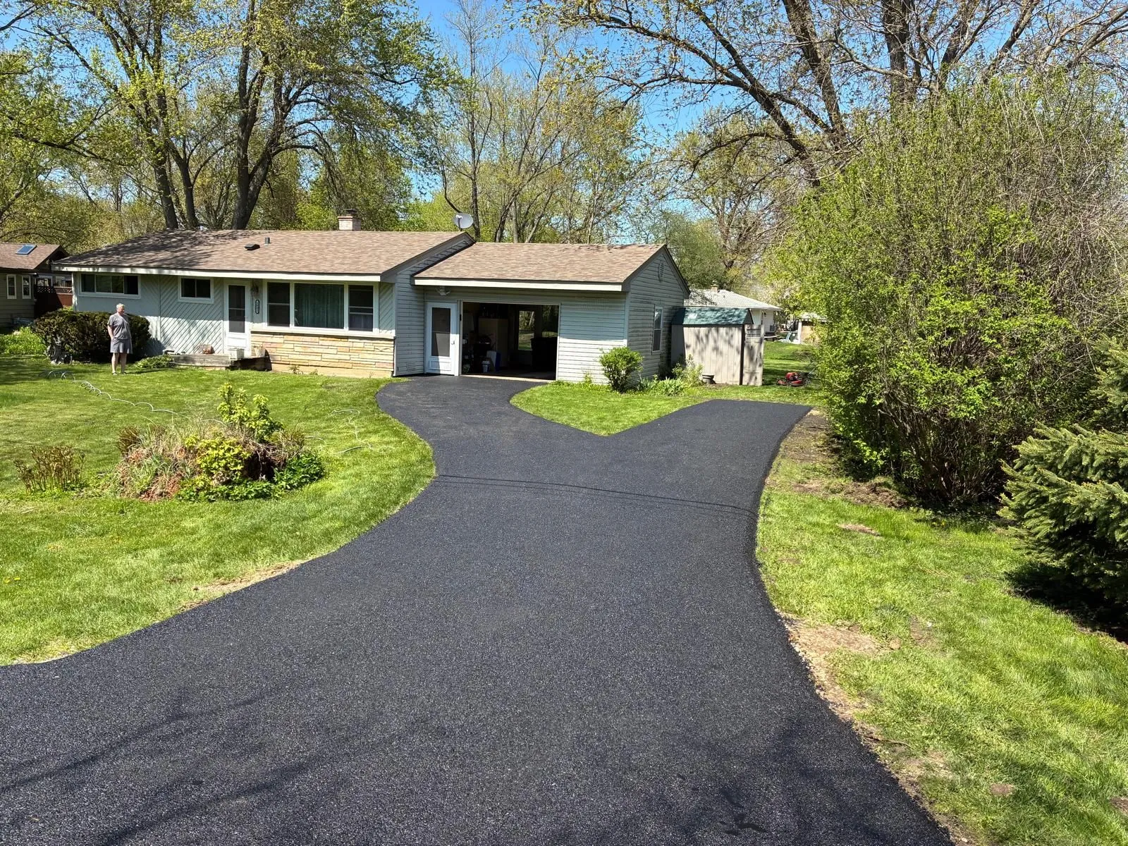 Single-story house with a curved black driveway, green lawn, and trees in the yard.