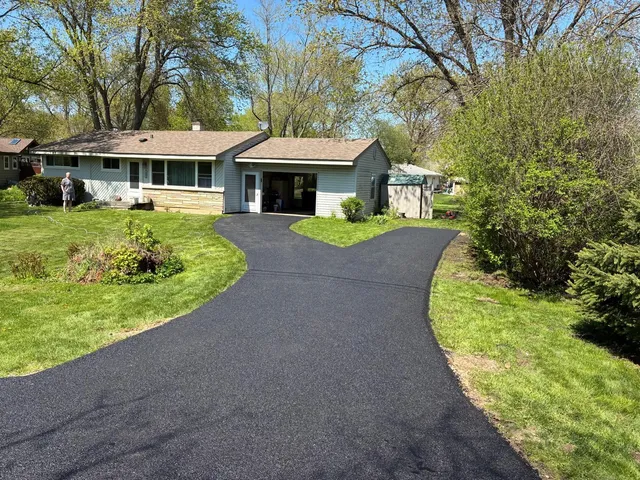 Single-story house with a curved black driveway, green lawn, and trees in the yard.