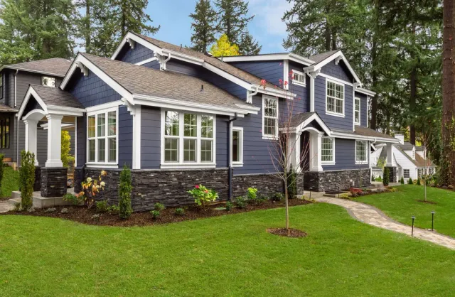 Large gray-blue house with white trim, stone foundation, and a green lawn surrounded by tall trees