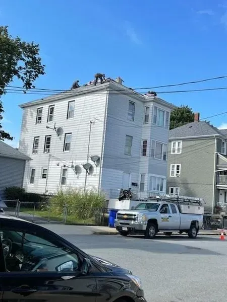 Workers on a roof, near a white building, blue sky, and a truck parked below.
