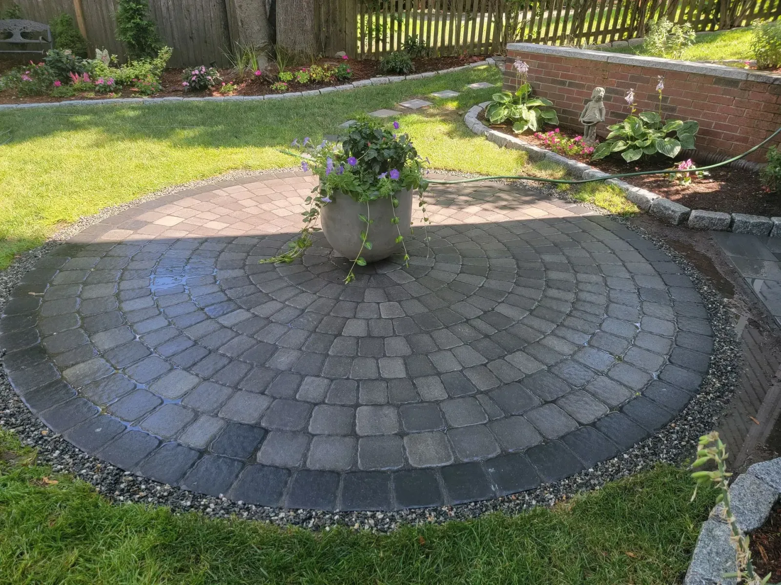 Circular paved patio with potted flowers, surrounded by grass and flower beds.