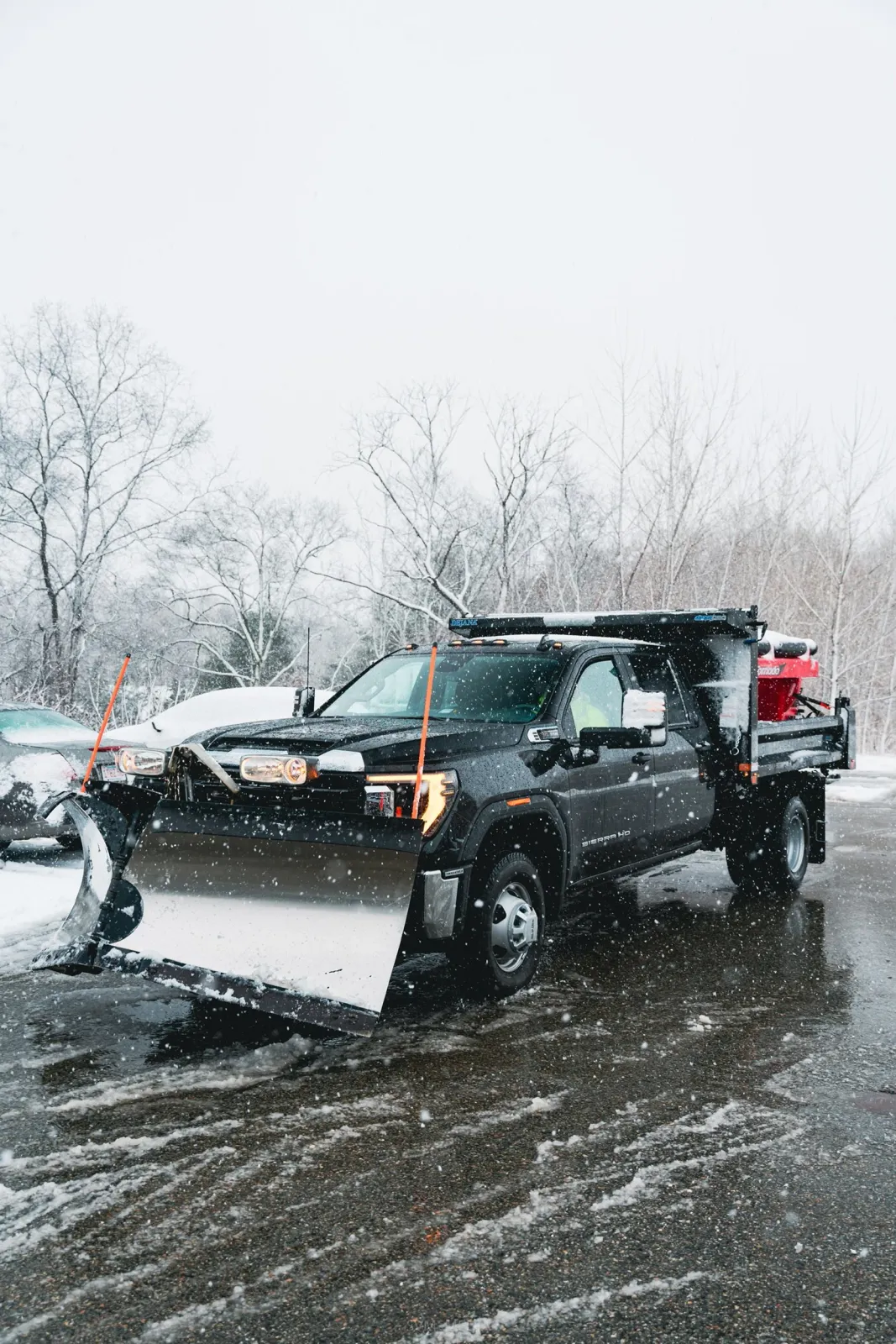 A black snow plow truck clearing snow from a parking lot on a snowy day, with leafless trees in the background.