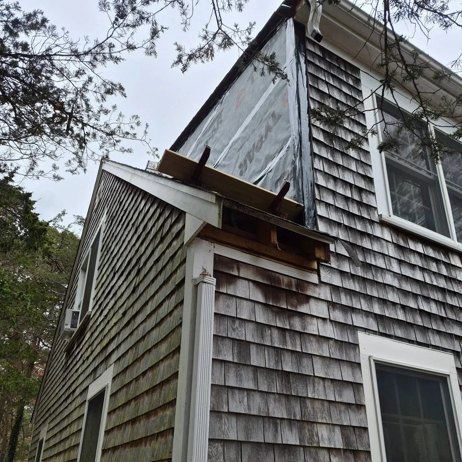 View of a corner of a house with weathered brown wooden shingles, a white gutter, and a window. Part of the house appears to be under renovation with a section covered in protective sheeting, and wooden supports holding up a small overhang or roof ex