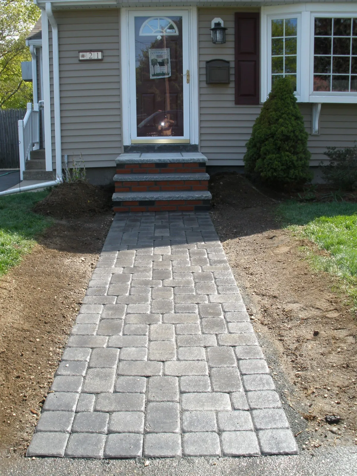 Stone walkway leading to the front door of a beige house with a small bush on the right.