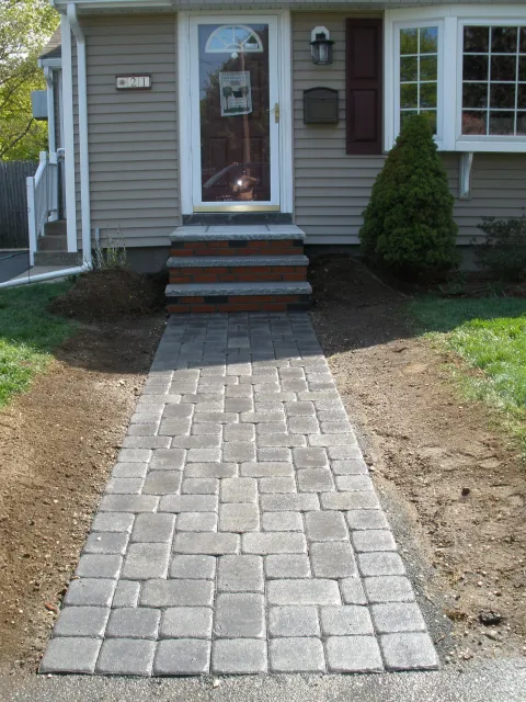 Stone walkway leading to the front door of a beige house with a small bush on the right.