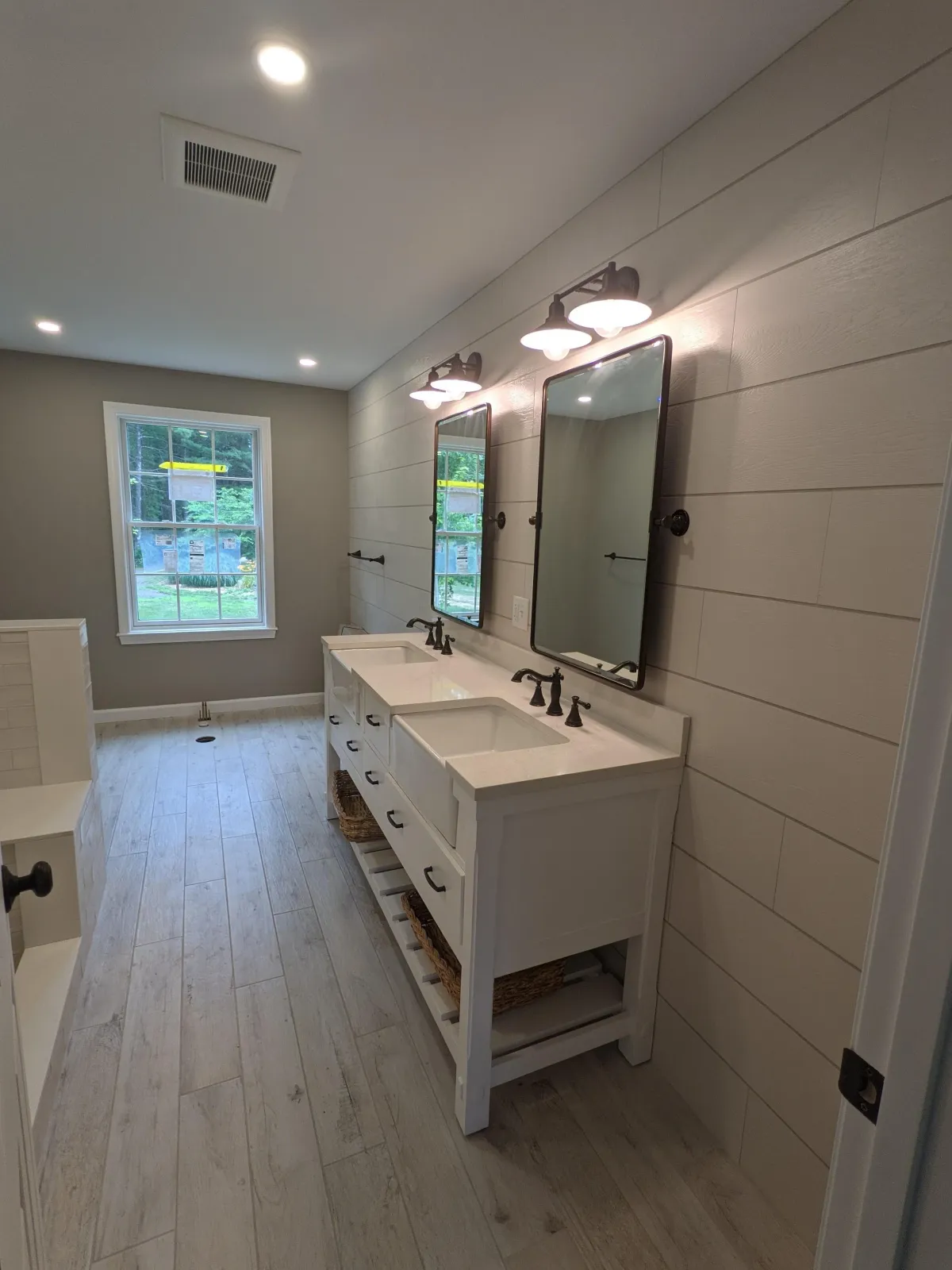 Modern bathroom with double vanity, white countertops, two black framed mirrors, black fixtures, three black vanity lights, light wood flooring, window.