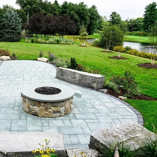 Custom stone fire pit and paved patio installed by Artisan Grounds in Greenland, NH, surrounded by lush landscaping with a natural pond and wooded backdrop.