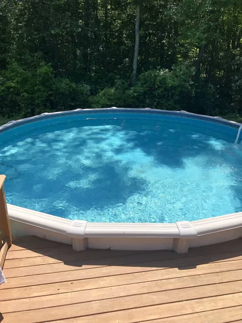 Above-ground swimming pool with clear water on a sunny day surrounded by a wooden deck and trees in the background.
