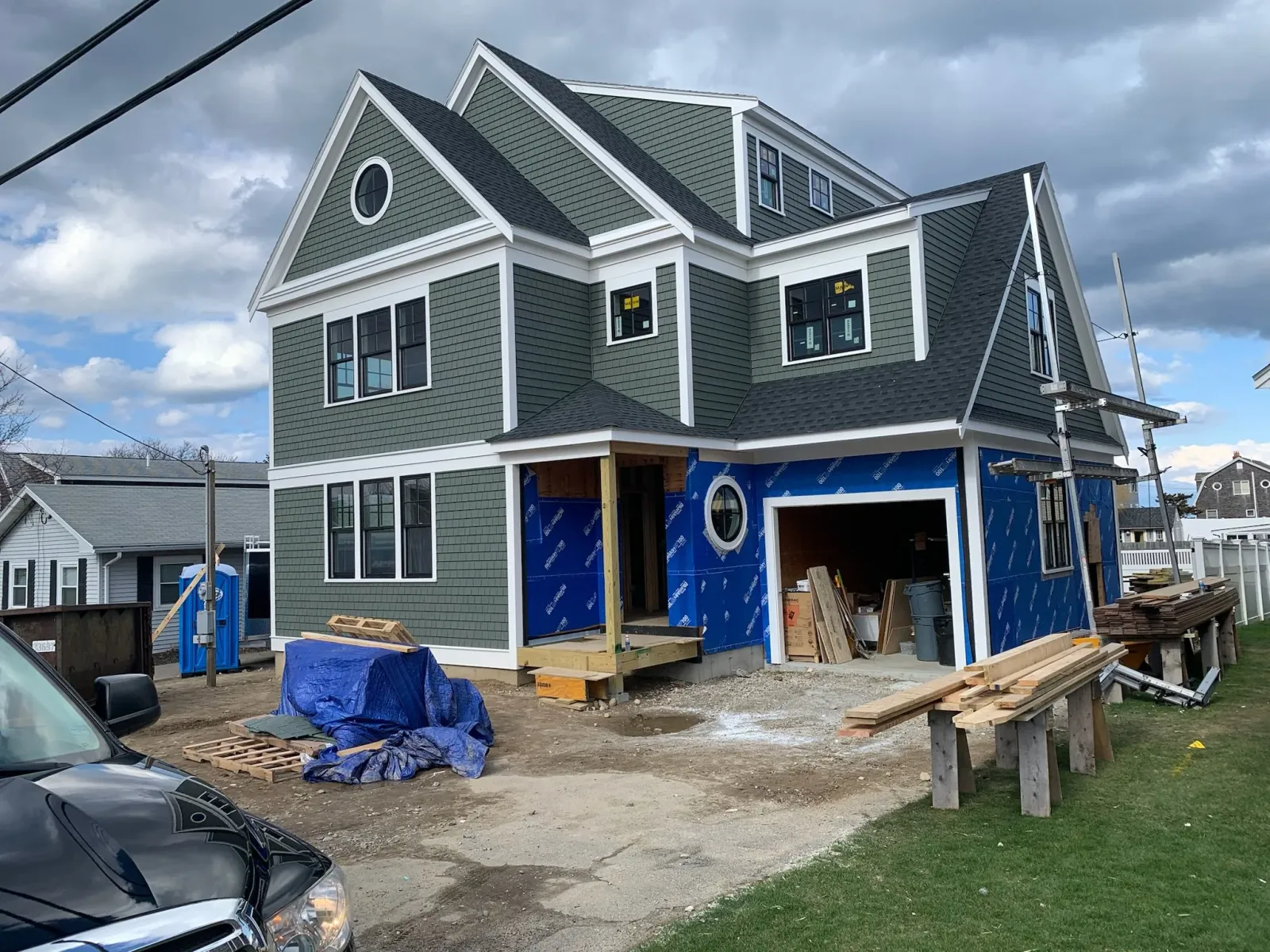 A multi-story house under construction with green siding, black shutters, and a garage. Construction materials and equipment are scattered around the muddy driveway, and a portable toilet is nearby. The sky is partly cloudy. Scituate, Norwell.