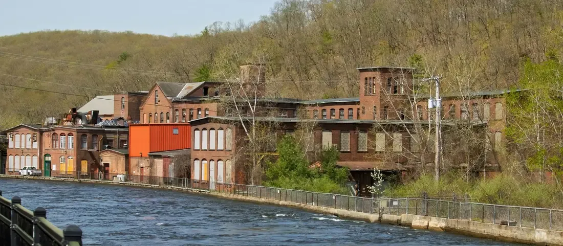 Old red brick mill buildings along a river below a wooded hillside