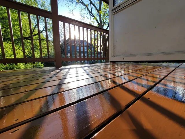 Close-up view of a wet wooden deck on Poplar Street, Roslindale with sunlight casting shadows from a railing, and trees and houses in the background.