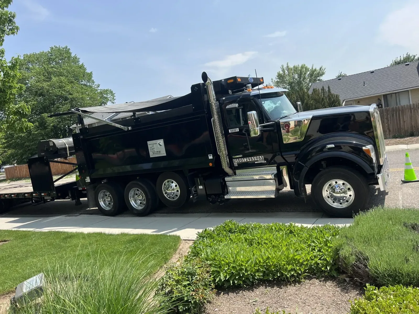 Black dump truck parked on a driveway, with a trailer, next to green bushes and grass, on a sunny day.
