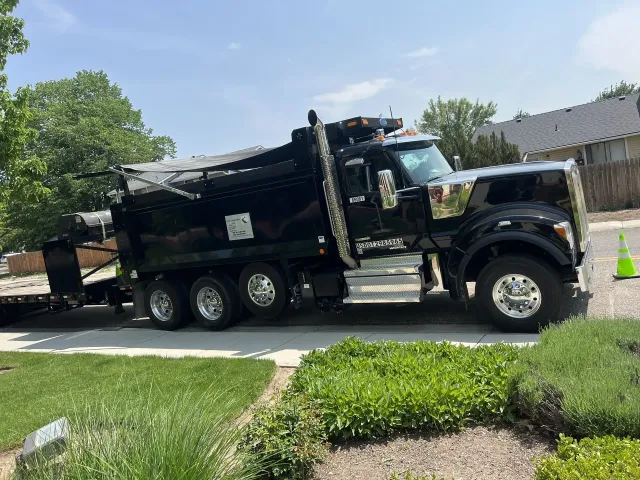 Black dump truck parked on a driveway, with a trailer, next to green bushes and grass, on a sunny day.