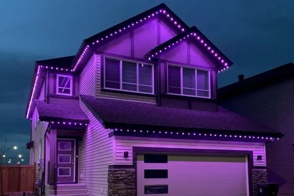 Suburban house with permanent lighting installed under the eaves, glowing a vibrant purple against a twilight sky. The lights accentuate the roofline and windows, creating a modern, festive look.