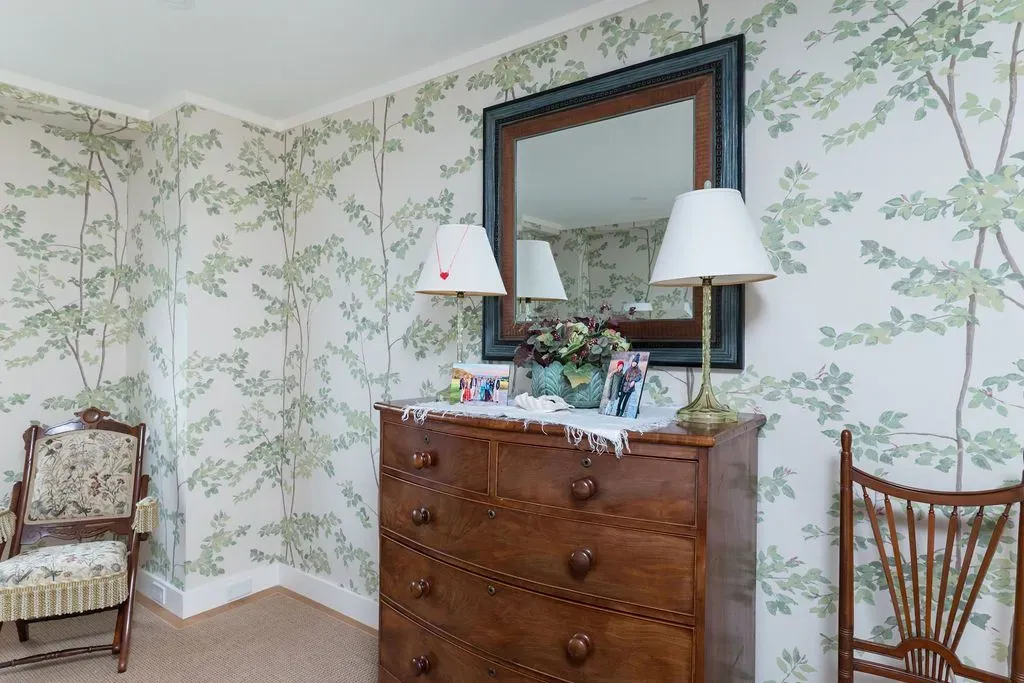 Floral wallpapered bedroom corner with wooden dresser, mirror, lamps, and two chairs