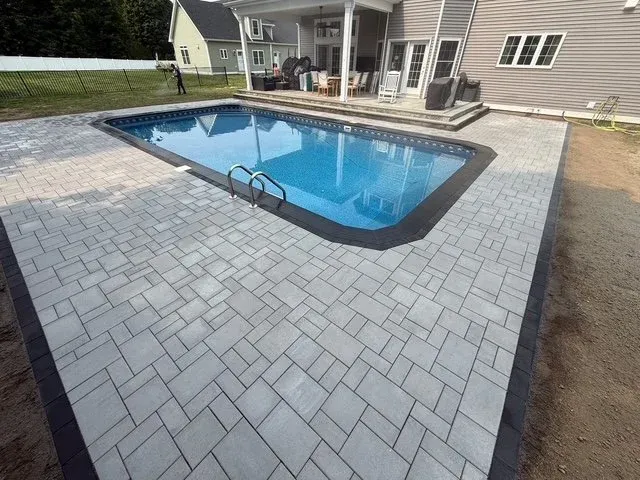 Backyard with a freeform swimming pool surrounded by new gray paver patio, adjacent to a house with sliding glass doors, outdoor seating, and a grassy yard with a white fence.