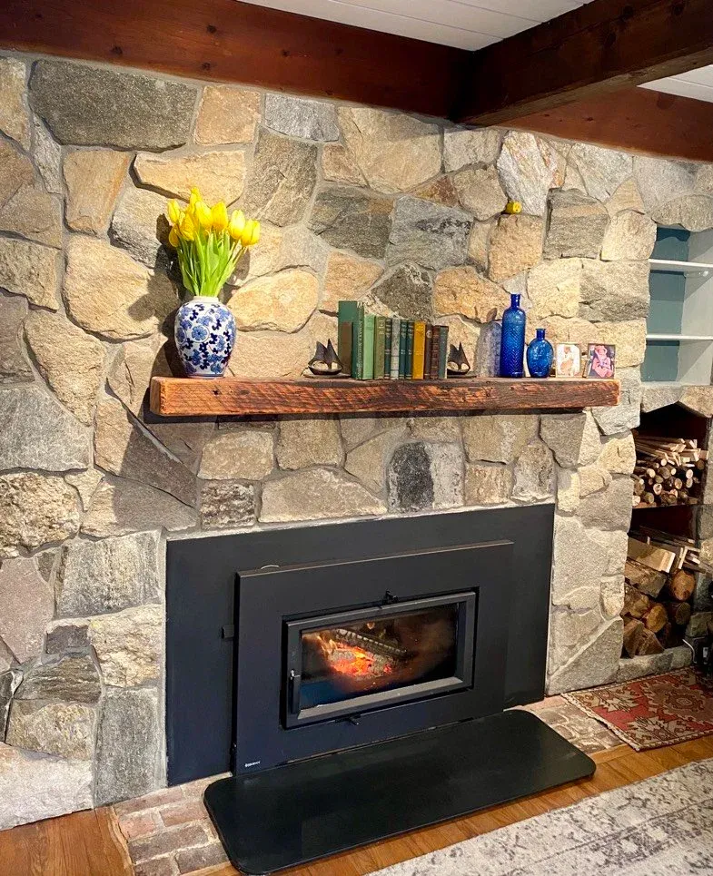 Mosaic stone fireplace with a custom wooden mantel, a vase of yellow tulips, books, decorative items, and firewood stacked to the side.
