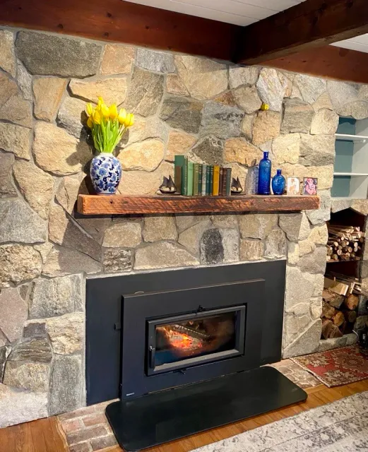 Mosaic stone fireplace with a custom wooden mantel, a vase of yellow tulips, books, decorative items, and firewood stacked to the side.