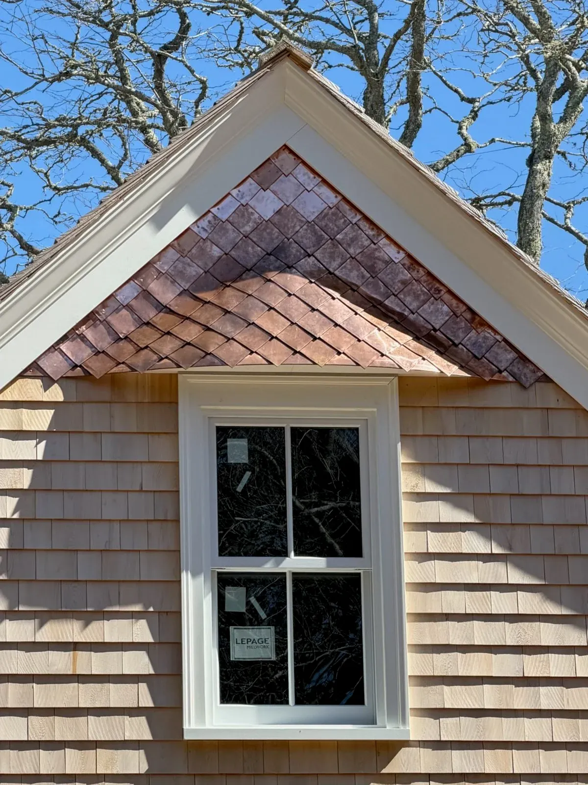 A house with beige wooden shingles, a white-framed window, and a roof with reddish-brown shingles, under a clear blue sky with leafless trees in the background.