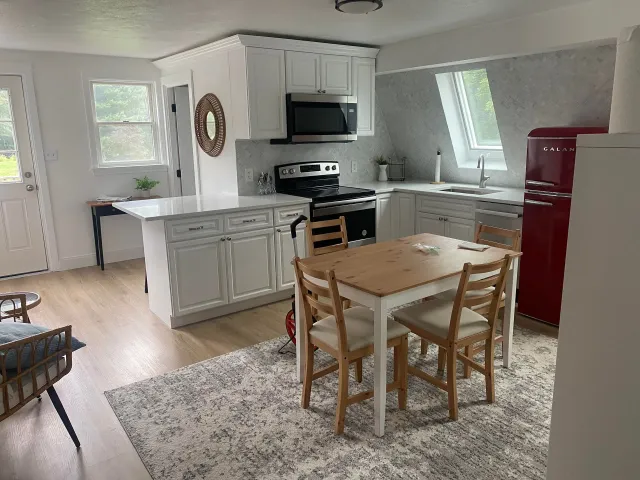 A bright kitchen with white cabinets, a light wood dining set, a red vintage-style refrigerator, and a patterned rug.