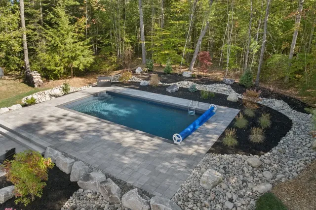 Rectangular in-ground pool surrounded by stone and plants, set in a wooded area with trees.