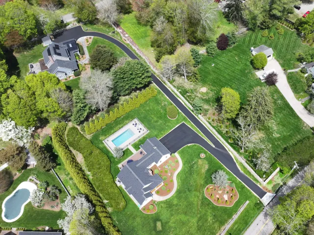 Aerial view of a suburban neighborhood with a curved driveway, dark-roofed house, manicured lawns and a rectangular backyard pool surrounded by trees.
