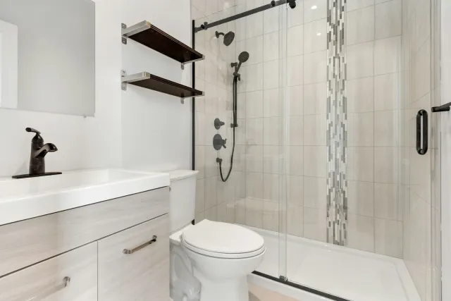 Modern bathroom with a white vanity, black faucet, wall-mounted shelves, toilet, and a shower with a glass door and black fixtures.