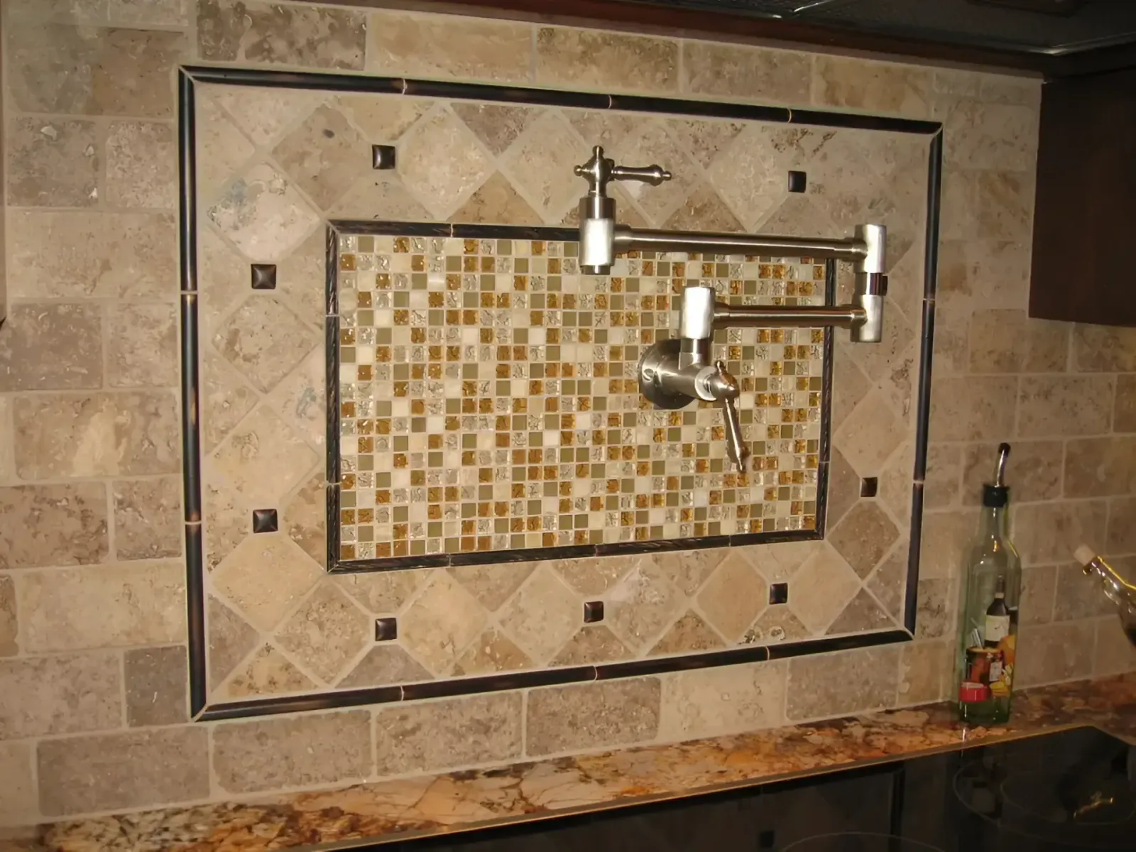 Tiled kitchen backsplash with a decorative mosaic panel and mounted metal spout above a stone countertop