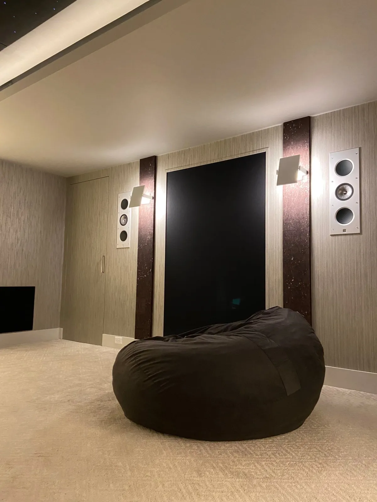 A room with beige carpet, a large black bean bag chair, wall-mounted speakers, and a large black panel on the wall.