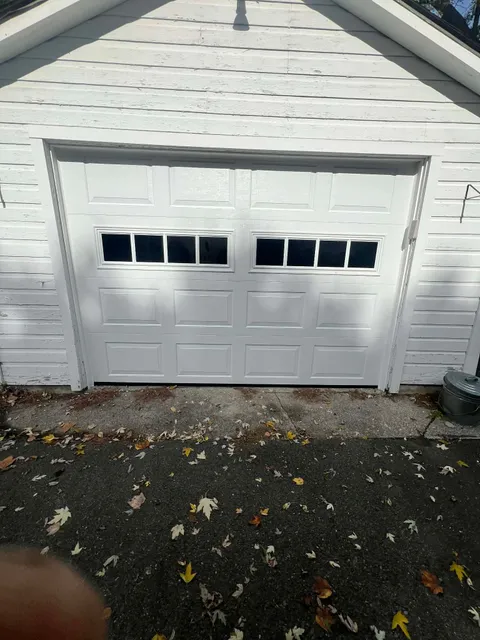 White garage door with windows, on a white building. Ground covered in leaves.