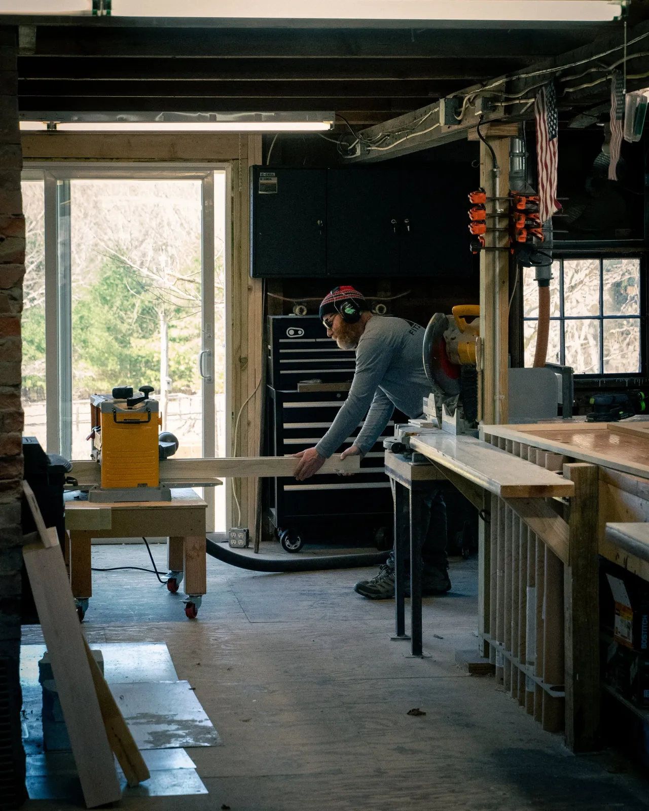 A person working in a woodworking shop, using a power saw on a piece of wood, wearing headphones and a beanie.