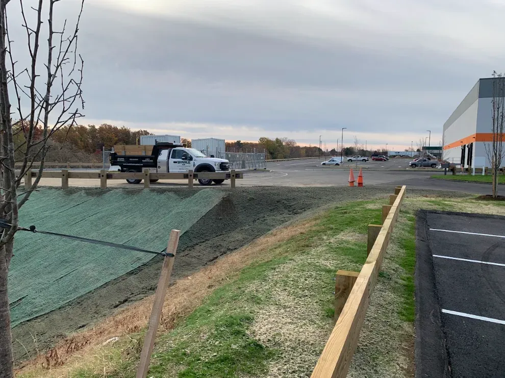 Construction site with trucks, gravel work area, orange cones, and a building beside a paved road.