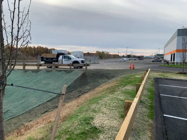 Construction site with trucks, gravel work area, orange cones, and a building beside a paved road.